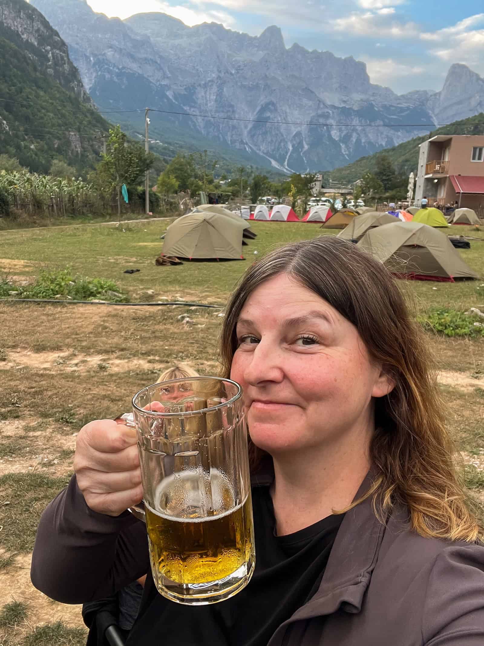 Sandra hält ein frisch gezapftes Bier in die Kamera, im Hintergrund Zelte und Berge in Theth