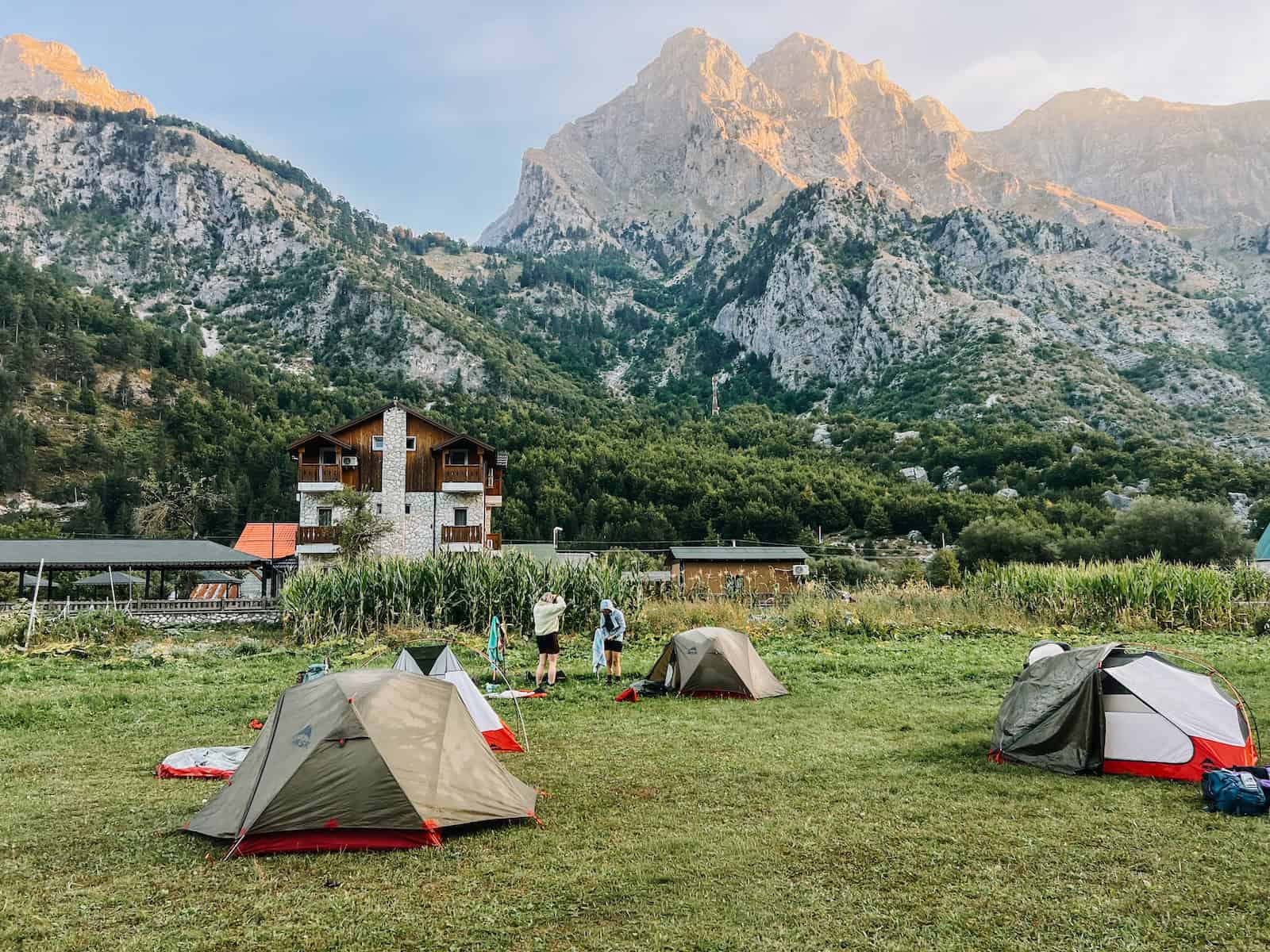 Zelte auf einer Wiese vor einem Berghaus, dahinter Berge im ersten Morgenlicht