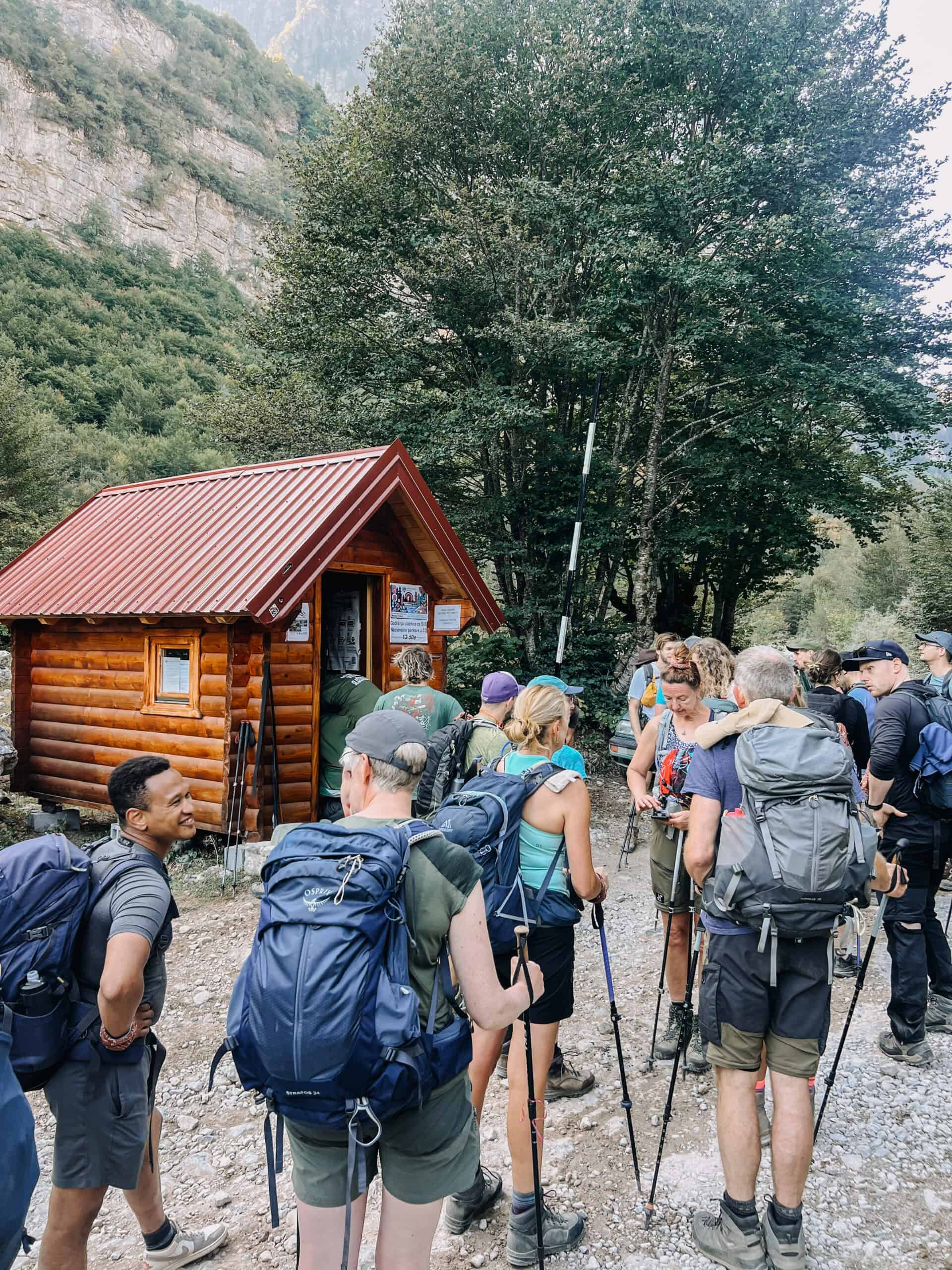 Wandergruppe mit Rucksäcken und Stöcken vor einer roten Holzhütte am Eingang des Nationalparks Prokletije