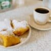 Drei Beignets mit Puderzucker auf einem Teller neben einer Tasse schwarzem Kaffee