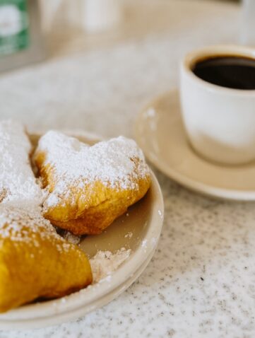 Drei Beignets mit Puderzucker auf einem Teller neben einer Tasse schwarzem Kaffee