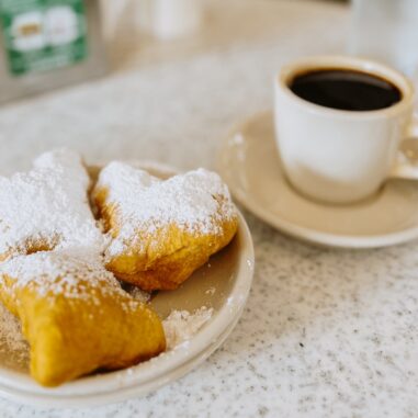 Drei Beignets mit Puderzucker auf einem Teller neben einer Tasse schwarzem Kaffee
